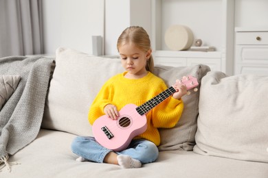 Photo of Little girl playing ukulele on sofa at home