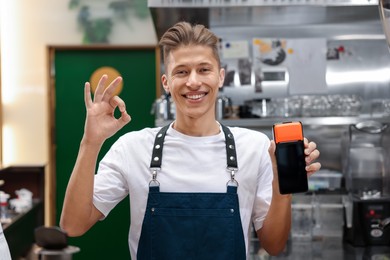 Photo of Smiling cafe worker with payment terminal showing OK gesture indoors