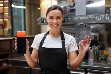 Photo of Smiling cafe worker with payment terminal showing OK gesture indoors