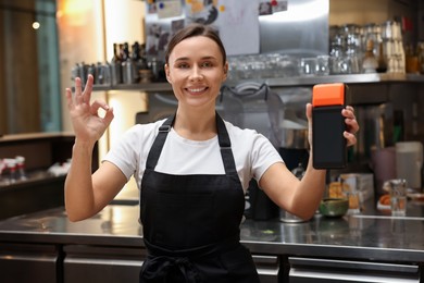 Photo of Smiling cafe worker with payment terminal showing OK gesture indoors