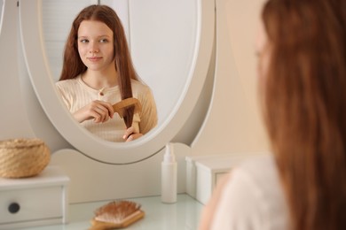 Photo of Beautiful teenage girl brushing her hair with comb near mirror at home, selective focus