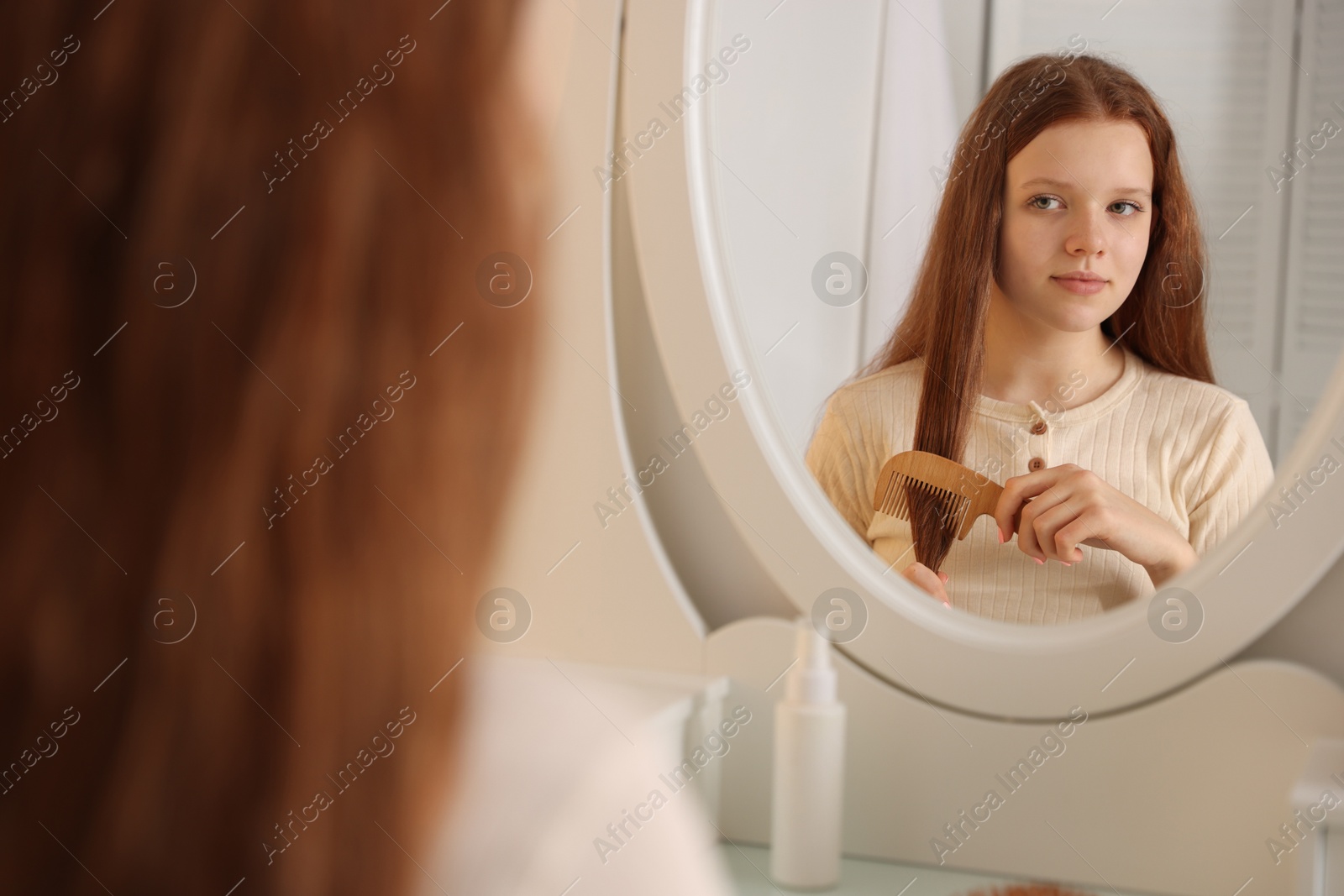 Photo of Beautiful teenage girl brushing her hair with comb near mirror at home, selective focus