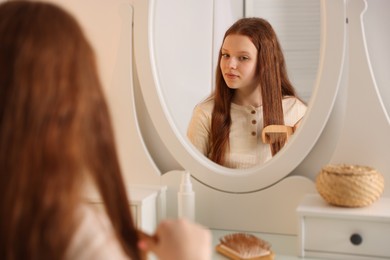 Photo of Beautiful teenage girl brushing her hair with comb near mirror at home, selective focus