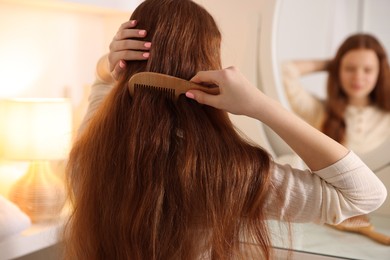 Photo of Teenage girl brushing her hair with comb near mirror at home, selective focus