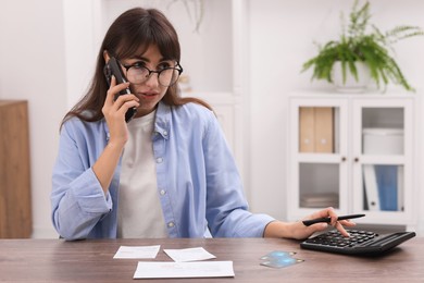 Photo of Paying bills. Woman with different invoices and calculator talking on phone at wooden table indoors