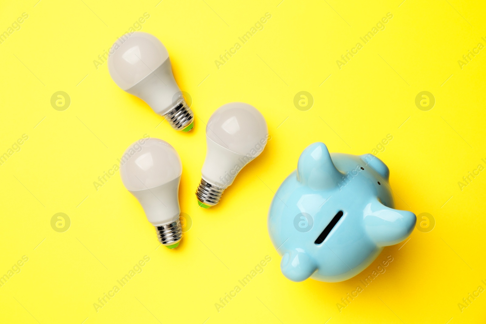 Photo of Piggy bank and light bulbs on yellow background, flat lay. Energy saving concept