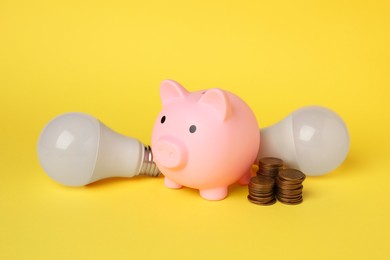 Photo of Piggy bank, coins and light bulbs on yellow background. Energy saving concept