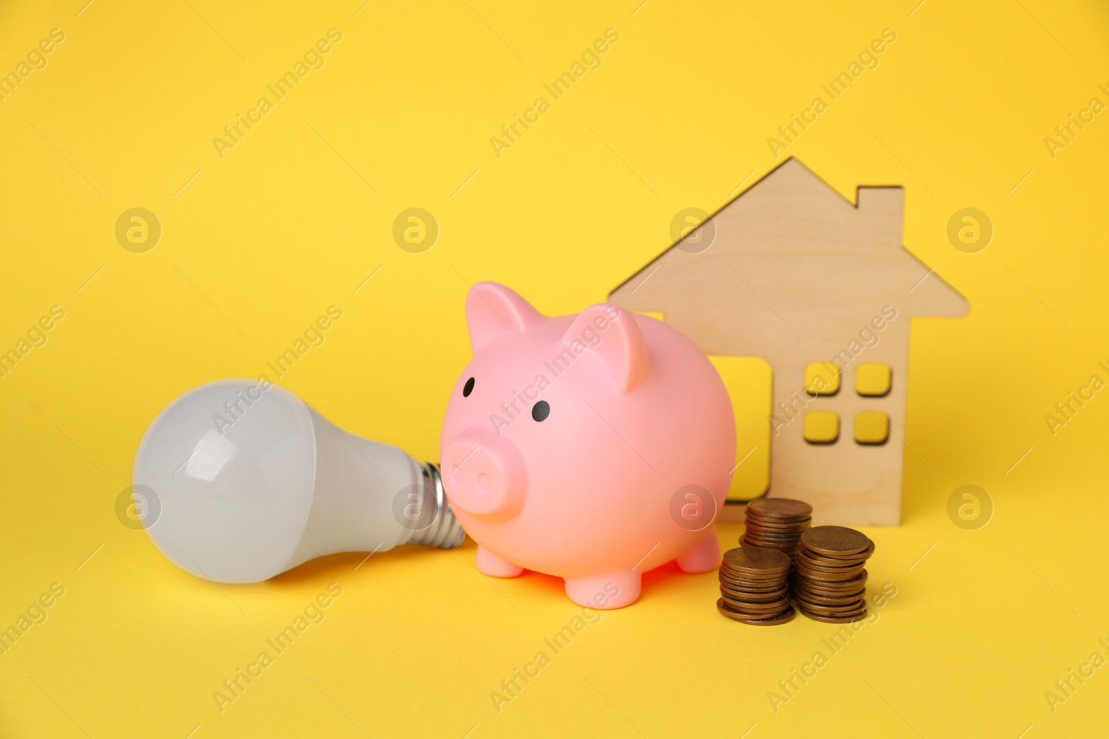 Photo of Piggy bank, coins, house model and light bulb on yellow background. Energy saving concept