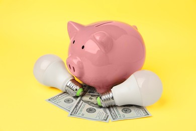 Photo of Piggy bank, dollar banknotes and light bulbs on yellow background, closeup. Energy saving concept