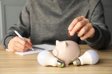 Photo of Man putting coin into piggy bank while taking notes at wooden table, closeup. Energy saving concept