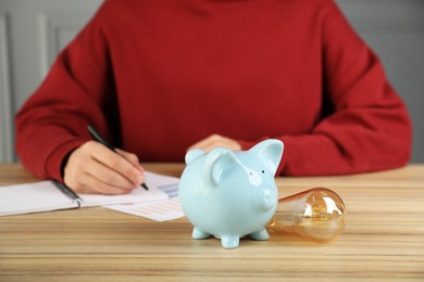 Photo of Man taking notes at wooden table, focus on piggy bank and light bulb. Energy saving concept