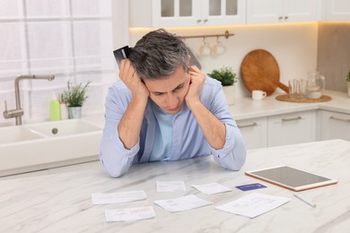 Photo of Paying bills. Confused man with different invoices, tablet and credit card at white marble table indoors