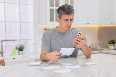 Photo of Man paying bills with smartphone at white marble table indoors