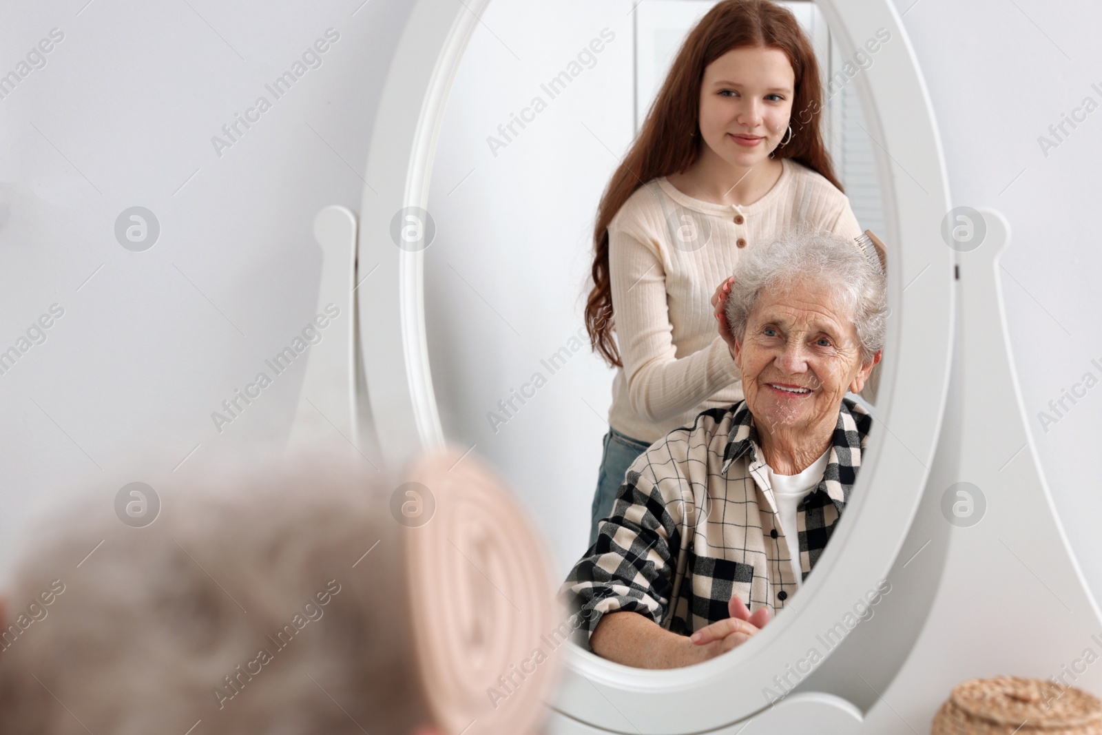 Photo of Granddaughter brushing her grandmother with brush near mirror at home