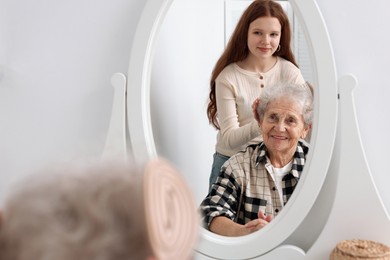 Photo of Granddaughter brushing her grandmother with brush near mirror at home