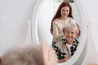 Photo of Granddaughter brushing her grandmother with brush near mirror at home