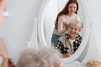 Photo of Granddaughter brushing her grandmother with brush near mirror at home