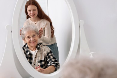 Photo of Granddaughter brushing her grandmother with brush near mirror at home