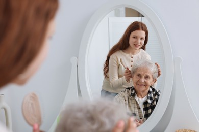 Photo of Granddaughter brushing her grandmother with brush near mirror at home