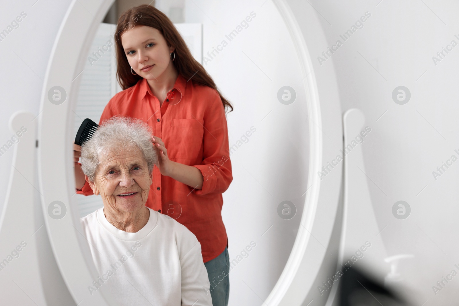 Photo of Granddaughter brushing her grandmother with comb near mirror at home