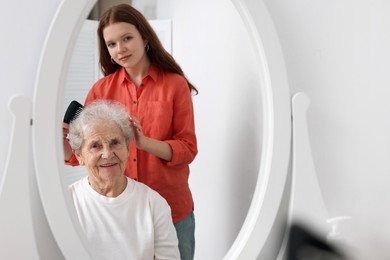 Photo of Granddaughter brushing her grandmother with comb near mirror at home