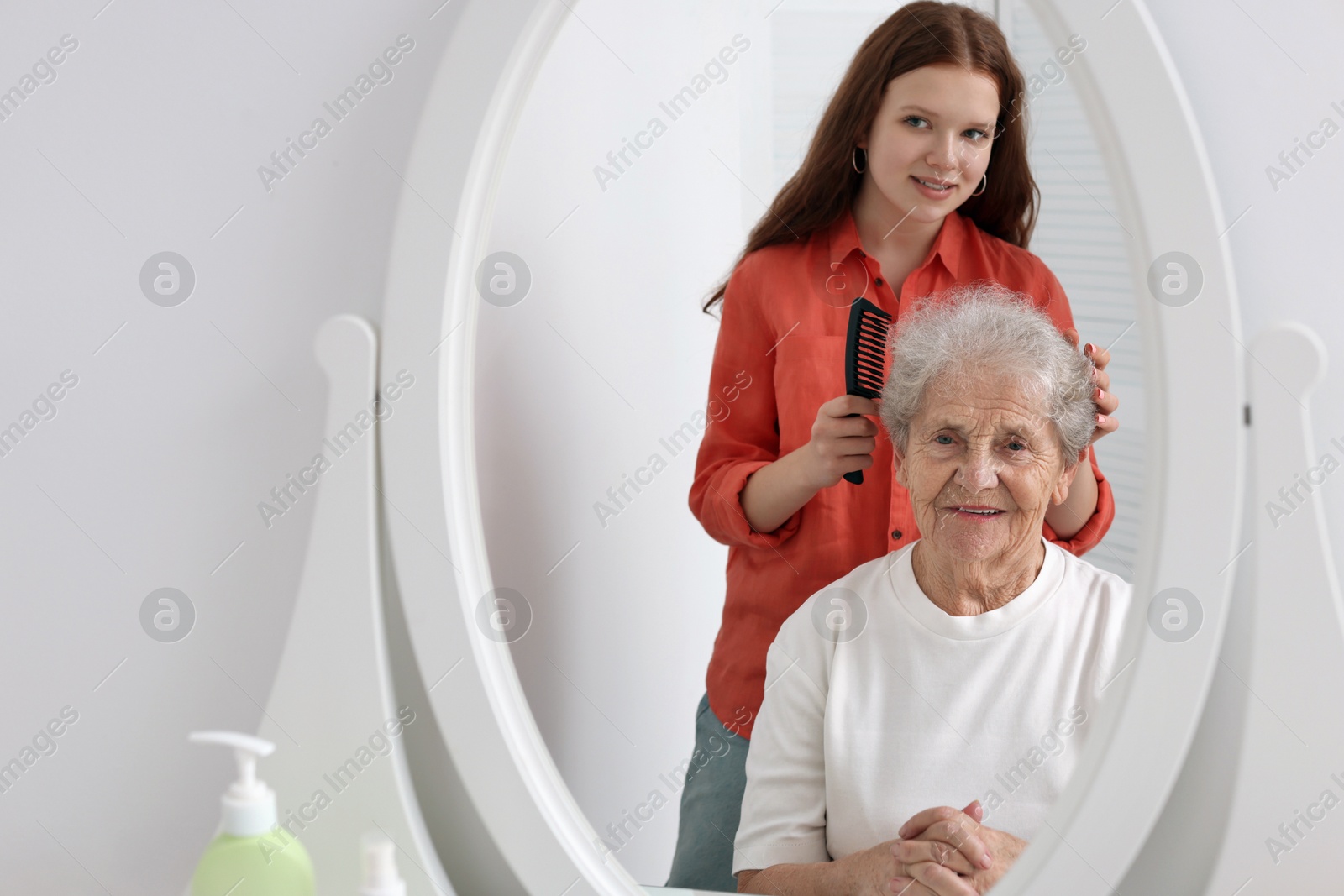 Photo of Granddaughter brushing her grandmother with comb near mirror at home