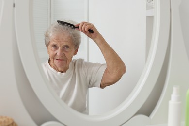 Photo of Senior woman brushing her hair with comb near mirror indoors