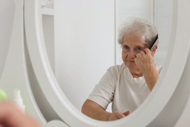 Photo of Senior woman brushing her hair with comb near mirror indoors