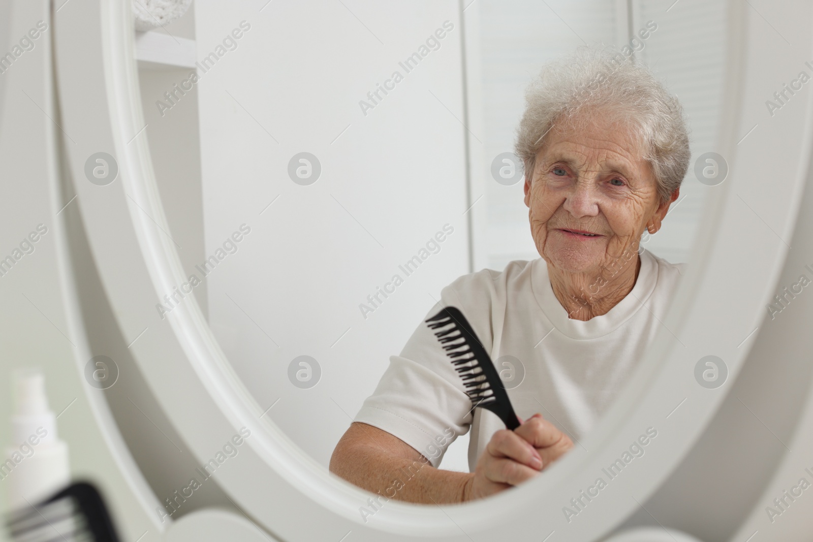 Photo of Senior woman brushing her hair with comb near mirror indoors