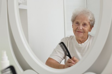 Photo of Senior woman brushing her hair with comb near mirror indoors