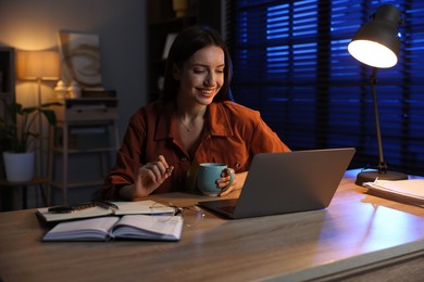 Photo of Woman with cup of coffee working on laptop at desk in home office