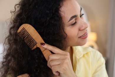 Photo of Smiling young woman brushing her curly hair with comb at home, closeup