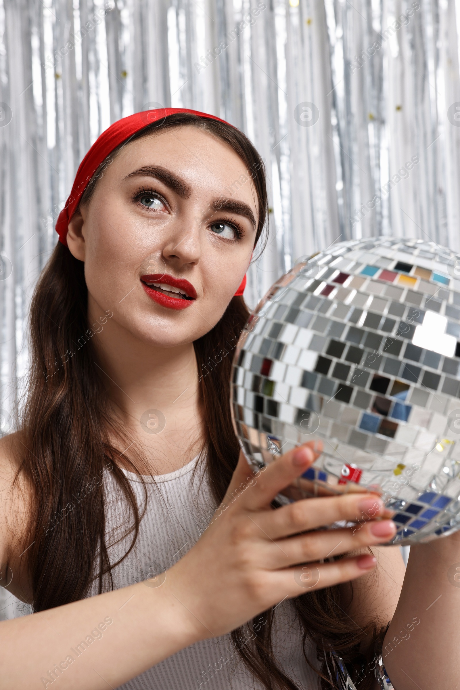 Photo of Portrait of beautiful young woman with disco ball against silver foil curtain