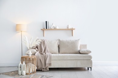 Photo of Comfortable sofa with pillows, blanket, lamp and different decor near white wall indoors