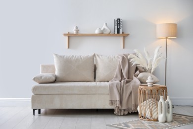 Photo of Comfortable sofa with pillows, blanket, lamp and different decor near white wall indoors