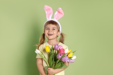 Photo of Happy little girl in headband with bunny ears holding bouquet of tulips on green background. Easter celebration