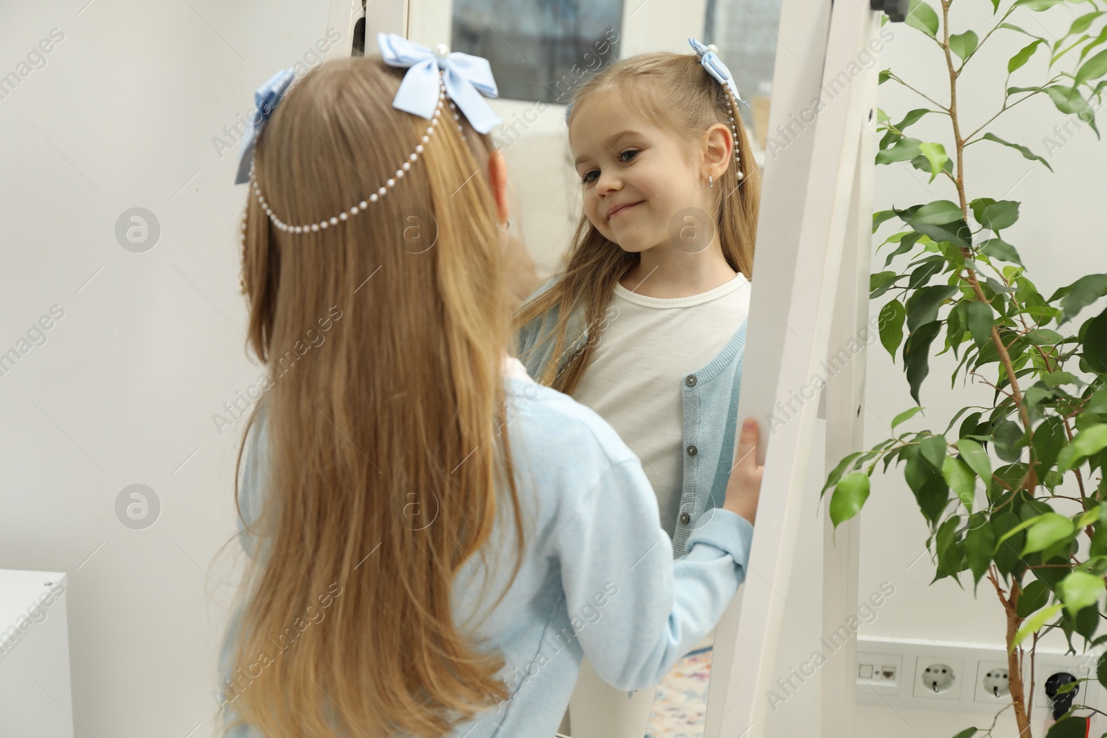 Photo of Cute little girl with beautiful hair accessory looking in mirror at home