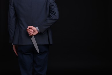 Photo of Businessman holding knife behind his back on black background, closeup. Space for text