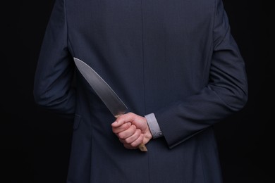 Photo of Businessman holding knife behind his back on black background, closeup