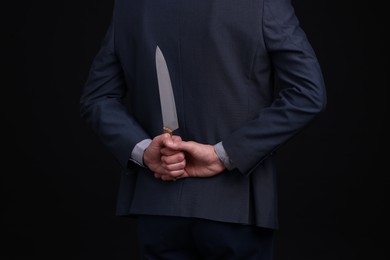 Photo of Businessman holding knife behind his back on black background, closeup