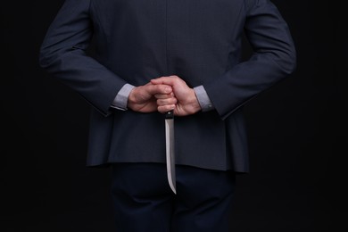 Photo of Businessman holding knife behind his back on black background, closeup