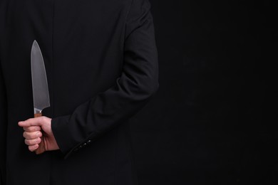 Photo of Businessman holding knife behind his back on black background, closeup. Space for text