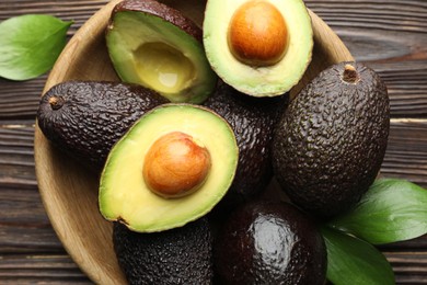 Photo of Whole, cut avocados and leaves in bowl on wooden table, flat lay