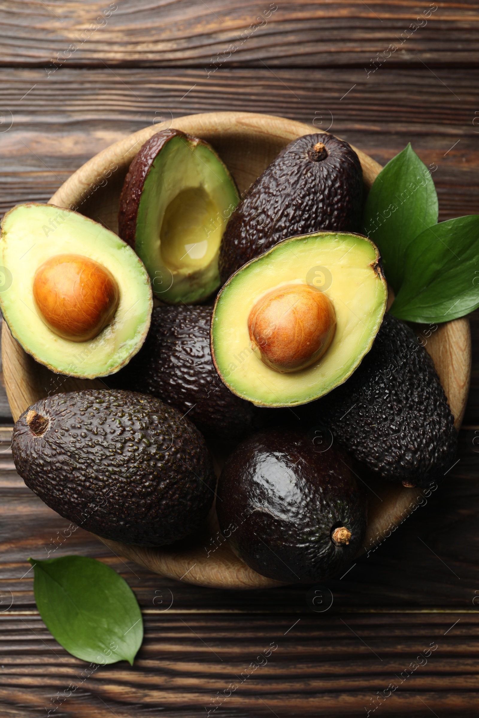 Photo of Whole, cut avocados and leaves in bowl on wooden table, flat lay