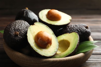 Photo of Whole, cut avocados and leaves in bowl on wooden table, closeup