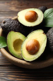 Photo of Whole, cut avocados and leaves in bowl on wooden table, closeup