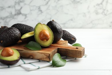 Photo of Whole, cut avocados and leaves on white wooden table against marble background, closeup. Space for text