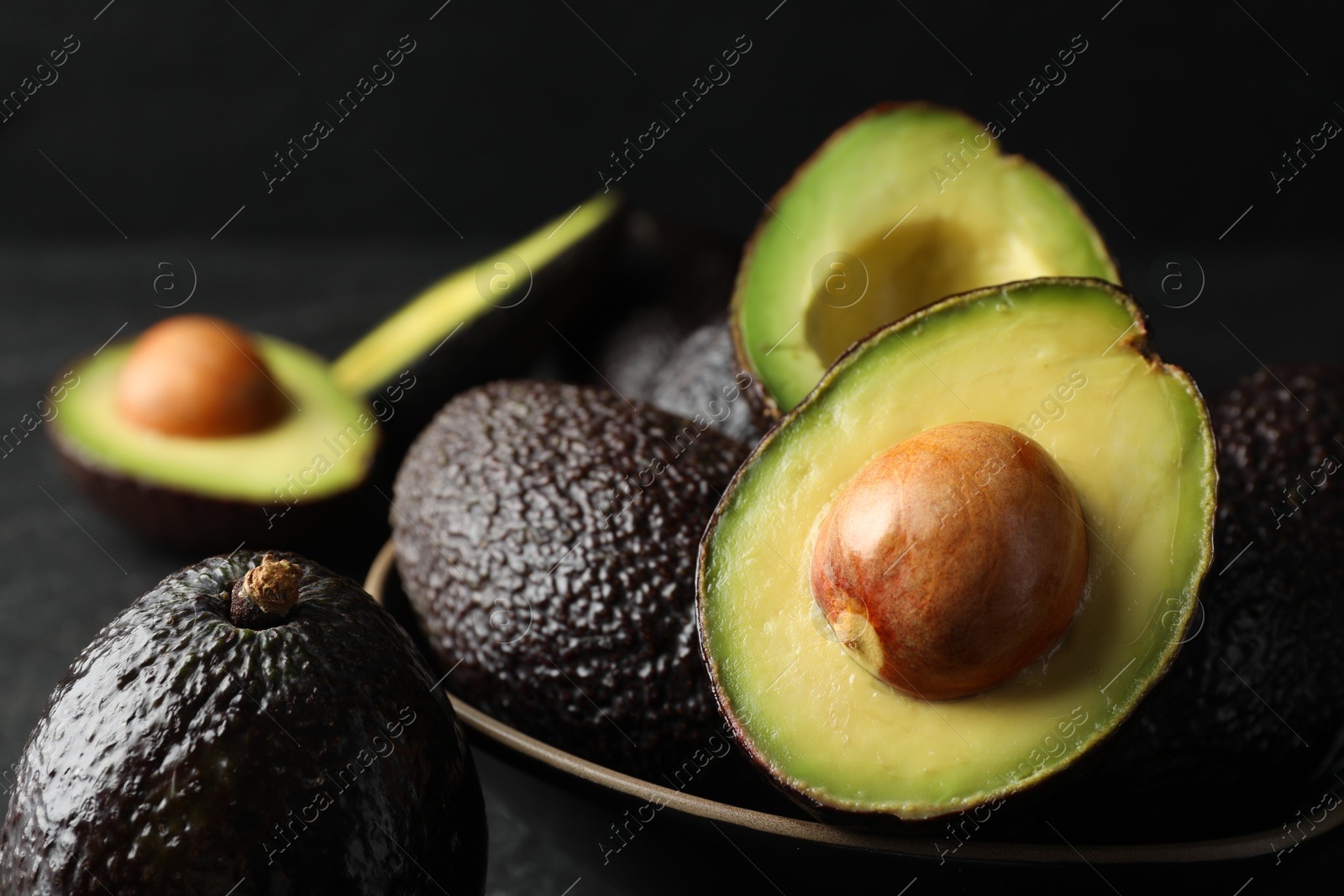 Photo of Whole and cut avocados on dark table against black background, closeup