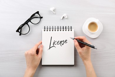 Image of Woman writing word License in notebook at white table, top view