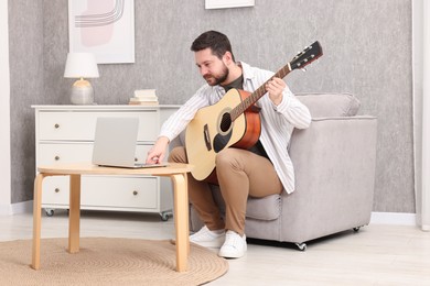 Photo of Relaxing hobby. Man playing guitar with online tutorial by laptop on armchair at home